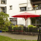 Das Bild zeigt den Garten des Seniorenzentrums Rothe Erde. Der Außenbereich ist mit einem roten Sonnenschirm über einem Tisch und Stühlen ausgestattet. Um den Tisch herum sind zahlreiche Pflanzen und Blumen zu sehen, die den Raum lebendig und einladend wirken lassen.