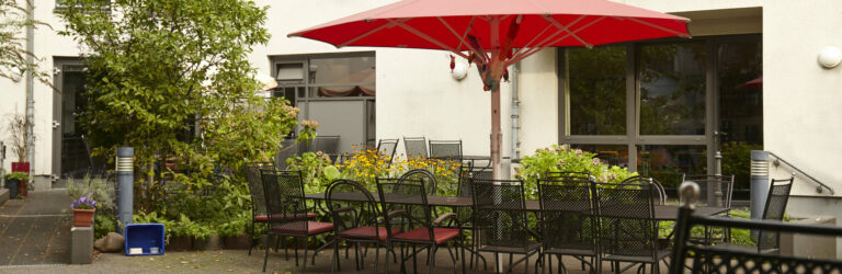 Das Bild zeigt den Garten des Seniorenzentrums Rothe Erde. Der Außenbereich ist mit einem roten Sonnenschirm über einem Tisch und Stühlen ausgestattet. Um den Tisch herum sind zahlreiche Pflanzen und Blumen zu sehen, die den Raum lebendig und einladend wirken lassen.