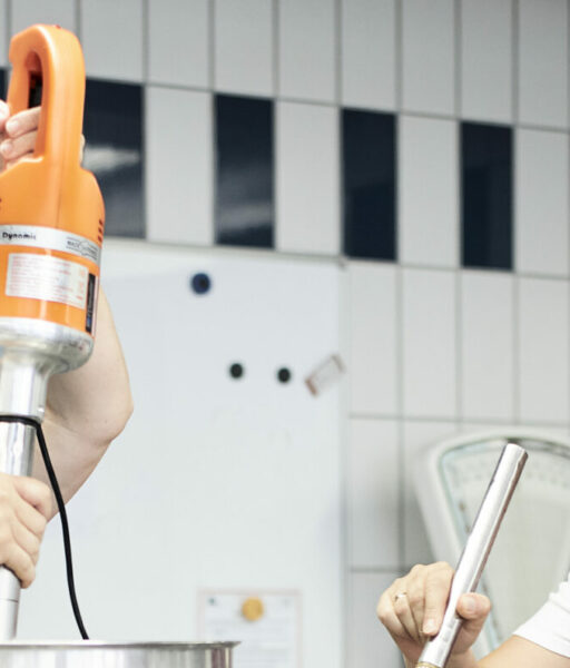 Isabella Plewnia und eine Mitarbeiterin arbeiten gemeinsam in einer Küche. Eine von ihnen bedient ein großes Küchengerät und beide lachen.