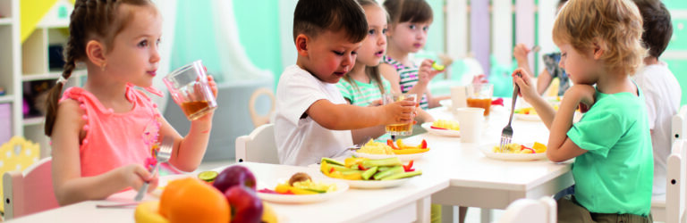 Mehrere Kinder sitzen an einem Tisch und essen Obst und Gemüse. Die Umgebung ist hell und einladend.